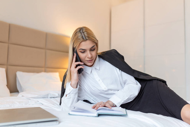 business, technology, internet and hotel concept - businesswomantalking on her mobile phone lying in hotel in bed and working late in hotel room, with her laptop computer, document and paperwork - Фото, изображение