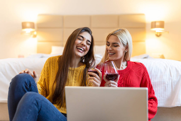 Twee aantrekkelijke meisjes, vrolijke beste vrienden die plezier hebben, films kijken op laptop en thuis rode wijn drinken. Twee glazen witte wijn in handen - Foto, afbeelding