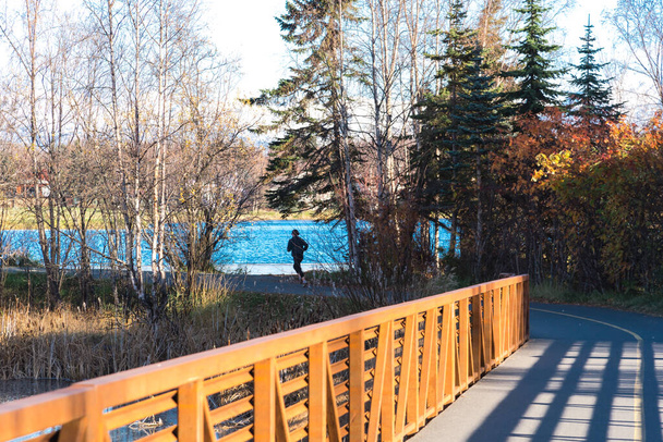 Küstenpfad an der Westchester Lagoon mit kaukasischer Dame, die am Seeufer entlang läuft und wunderschönem Herbstlaub in Anchorage, Alaska. Outdoor-Workout mit warmer Kleidung und Sportbekleidung, sonniger Tag - Foto, Bild