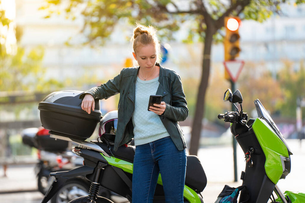 Портрет рыжеволосой женщины с помощью смартфона рядом со своим припаркованным мотоциклом - Фото, изображение