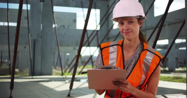 ハードハットと安全ベストの女性建設労働者は、クリップボードにメモを作成し、彼女の後ろの商業ビルフレームと現場のカメラで笑顔。4k - 映像、動画