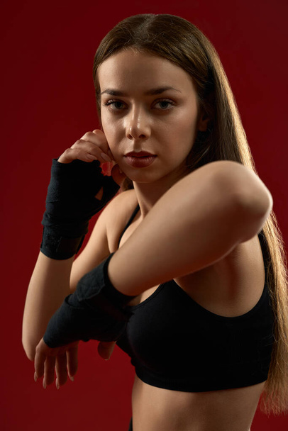 portrait of pretty brunette woman training I black sport bandages in studio. Beautiful sporty female trainer showing kick exercise on red background, with go hands. Concept of martial arts. - Foto, immagini
