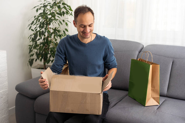 Handsome young man opening box with parcel while sitting on sofa at home - Foto, immagini