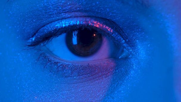 Womens eye with shiny make up with sparkles and arrow looking straight. Look of female eye with eyelashes and eyebrows illuminated by blue neon light. Macro portrait of female eye with brown iris - Fotoğraf, Görsel