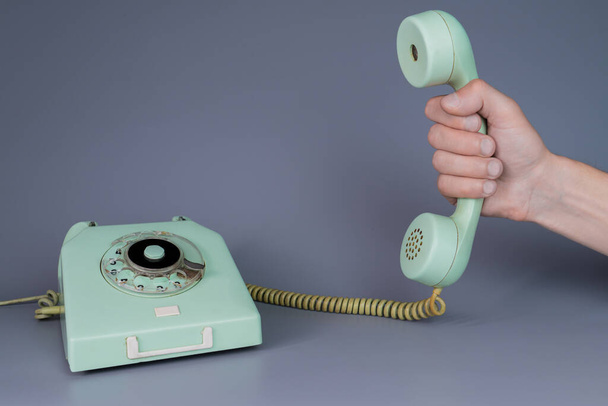 Male hand holding an old blue plastic telephone receiver near rotary telephone on gray background. Close up remote handset from retro home phone apparatus in the hands of man. Concept of communication - Foto, immagini