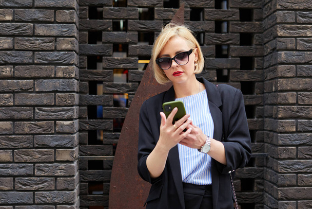 Side view of concentrated woman in stylish outfit text messaging on mobile phone while walking along pavement near contemporary building - Photo, Image