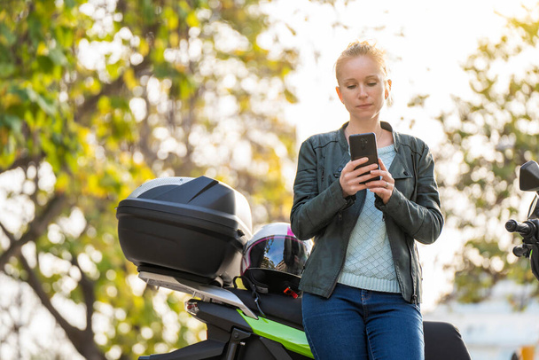 Portrét rudovlasé ženy pomocí smartphonu vedle její zaparkované motocyklu - Fotografie, Obrázek