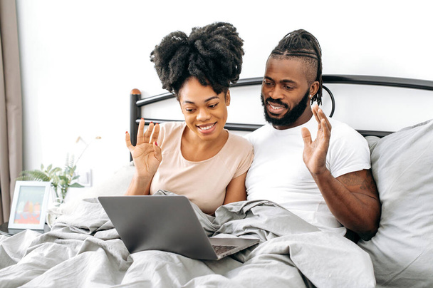 Comunicazione video a distanza. Positivi sposi afroamericani felici, marito e moglie, sono seduti in un letto in camera da letto, utilizzando il computer portatile, parlando in videoconferenza con amici o familiari, sorridendo. - Foto, immagini