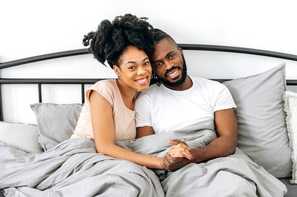 Tatmin olmuş pozitif eşlerin fotoğrafı, Afro-Amerikan erkek ve kadın, ev kıyafetleri içinde, yatak odasında rahat bir yatakta oturuyor, birbirlerine sarılıyor, kameraya bakıyor, gülümsüyor, mutlu hissediyorlar. - Fotoğraf, Görsel