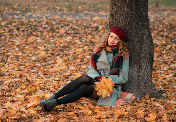 Kręcone wesołe dziewczyny z pięknymi oczami noszące modny szary płaszcz i szary szalik siedzi w pobliżu drzewa w parku. Jesienią. Portret. - Zdjęcie, obraz