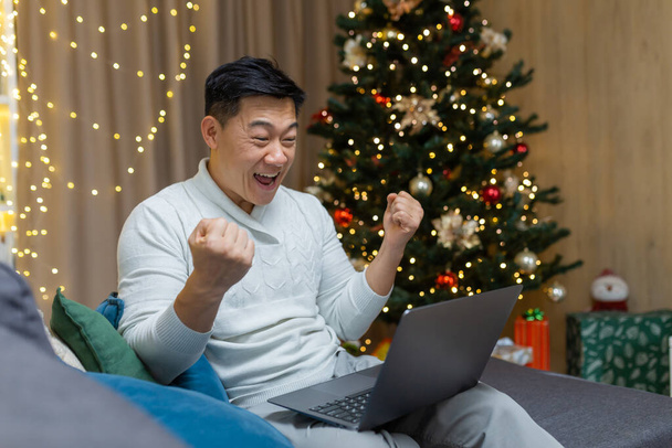 A young Asian man is playing games on a laptop, sitting at home on the couch near the nine-year-old Christmas tree. Happy with success, makes yes with his hands. - Valokuva, kuva
