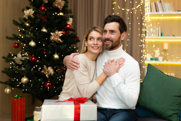 Portrait of a happy young couple, family. Young man and woman celebrating Christmas holidays at home. They sit on the sofa near the Christmas tree with gifts, hug, rejoice, look away. - Photo, image