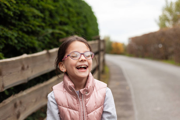 Little piękne słodkie dziewczyny w okularach śmiejąc się w pobliżu drewnianego ogrodzenia. Wiejskie dziecko w kamizelce na spacerze po wsi - Zdjęcie, obraz