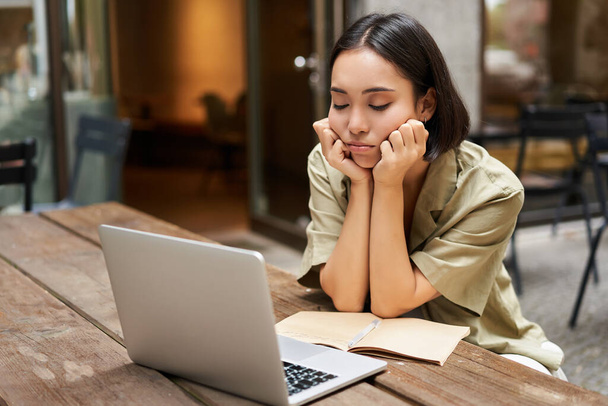 Znudzona dziewczyna patrzy na laptopa, siedzi na zewnątrz w kawiarni, słucha nudnych spotkań online, pracuje i jest smutna. - Zdjęcie, obraz
