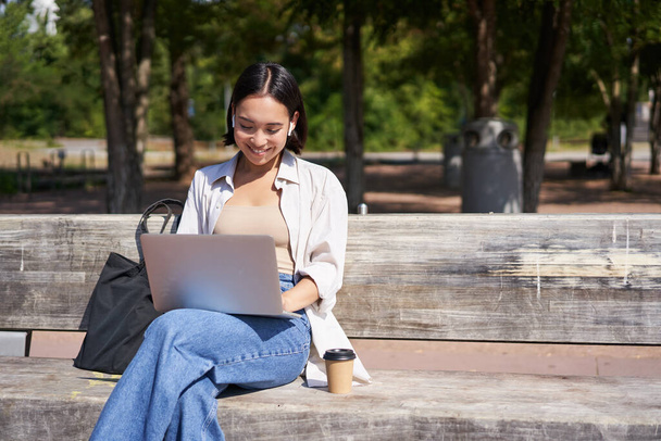 Retrato de la mujer coreana sentada con el ordenador portátil en el parque en el banco, trabajando al aire libre, estudiante haciendo la tarea, beber café para llevar. - Foto, imagen