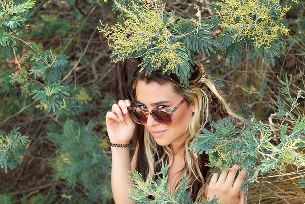 A young white woman in sunglasses looking at the camera standing among the branches of a tree on a sunny day - Fotoğraf, Görsel