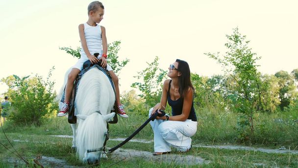 A woman and a boy are walking around the field, son is riding a pony, mother is holding a pony for a bridle. Cheerful, happy family vacation. Outdoors, in summer, near the forest. High quality photo - Fotoğraf, Görsel
