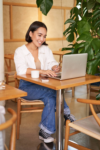Stylowa bizneswoman z laptopem, siedzi w kawiarni i pracuje na komputerze, zarządza biznesem i pije kawę. - Zdjęcie, obraz