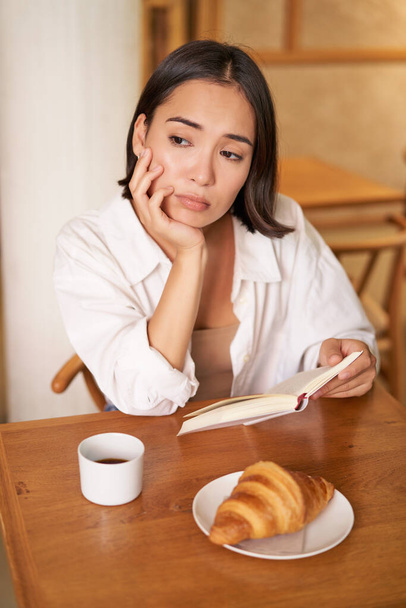 Vertical shot de chica asiática se sienta solo en la cafetería, lee libro y se ve molesto, bebe café con croissant. - Foto, Imagen