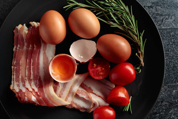 Uzená slanina s vejci, rajčaty a rozmarýnem na černém talíři. Horní pohled.  - Fotografie, Obrázek