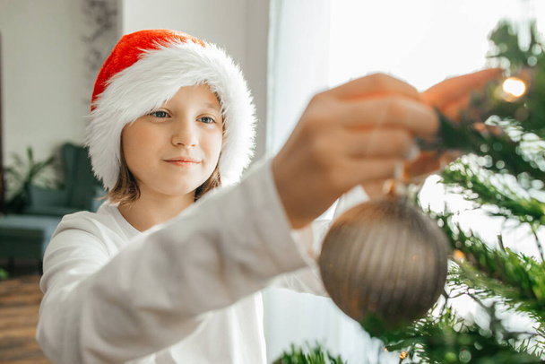 Ευτυχισμένη έφηβη διακοσμεί το χριστουγεννιάτικο δέντρο στο σπίτι την παραμονή των εορτών. Τα παιδιά περιμένουν τα Χριστούγεννα και την Πρωτοχρονιά.  - Φωτογραφία, εικόνα