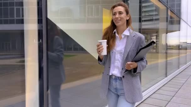 Sikeres fiatal gyönyörű nő táblagéppel és kávéscsészével sétál az üzleti épületek között, és örömmel mosolyog. Vonzó üzletasszony. Karrieresek. Kiváló minőségű 4k felvételek - Felvétel, videó
