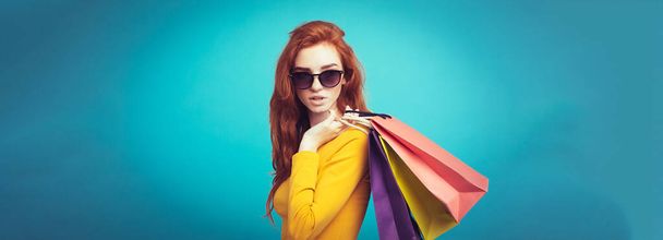 ショッピングのコンセプト - ショッピング バッグとカメラを見て笑っている肖像若い美しい魅力的な redhair 女の子をクローズ アップ。青いパステル背景。コピー スペース. - 写真・画像