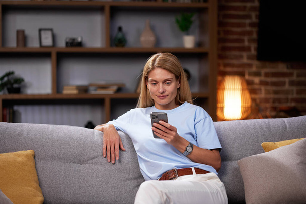 Blonde girl sitting on the couch looks into the smartphone, chatting, relaxing at home. Smiling adult woman communicates friendly by typing a message on the phone - Photo, image