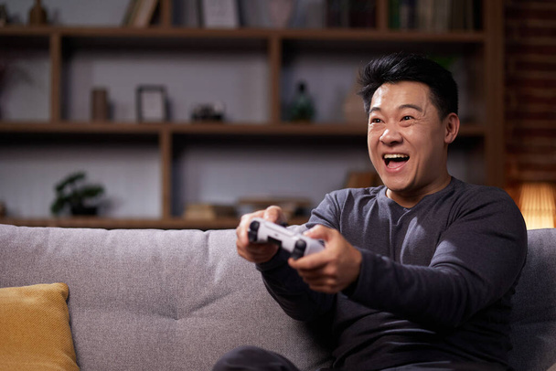 Happy Asian man using a wireless joystick and playing video games at home. Joyful man using PlayStation and competing in virtual games while resting on the sofa. - Foto, Imagen