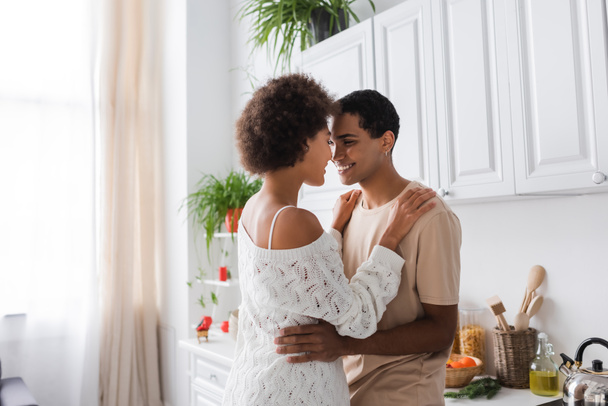 curly african american woman in white openwork sweater smiling near happy boyfriend in kitchen - Photo, Image
