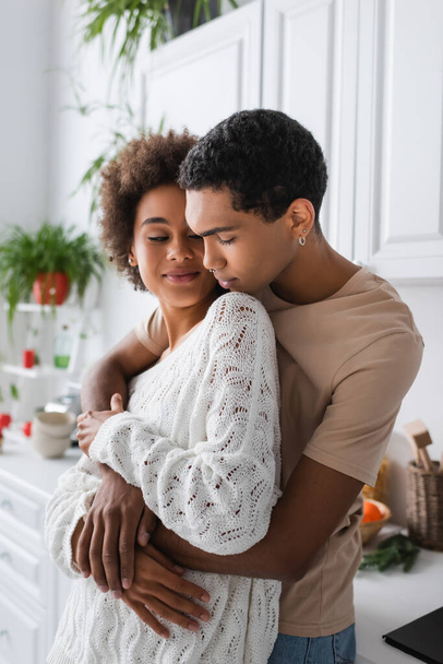 young african american man with piercing embracing smiling woman in white openwork sweater in kitchen - Photo, image