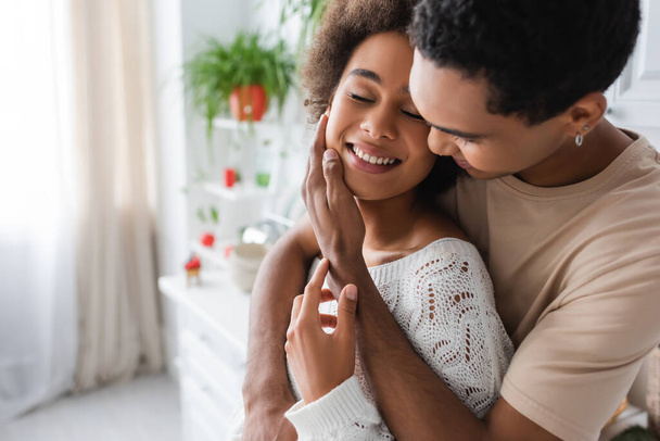 young african american man hugging and touching face of happy woman with closed eyes in kitchen - Foto, imagen