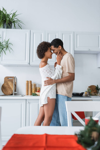 boční pohled na africké americké ženy v bílém svetru objímání s přítelem v kuchyni - Fotografie, Obrázek