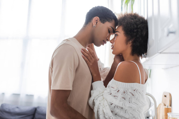 side view of african american man in t-shirt and sexy woman in knitted sweater standing face to face in kitchen - Foto, immagini