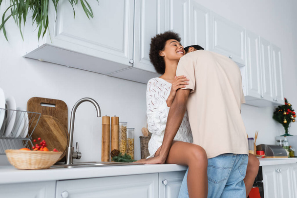 africano americano hombre abrazando feliz novia en blanco suéter sentado en worktop en cocina - Foto, Imagen
