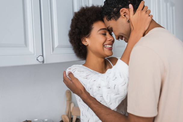 cheerful african american woman with closed eyes hugging boyfriend near white furniture in kitchen - Zdjęcie, obraz