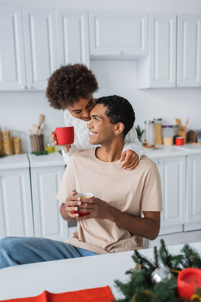 curly african american woman with red cup near smiling boyfriend in kitchen - Photo, Image