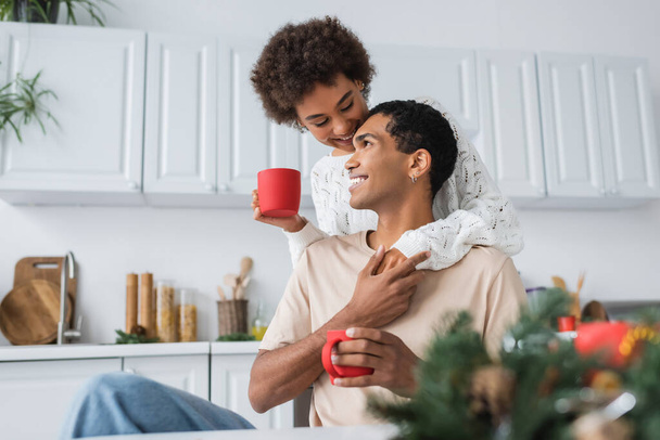 afro-américaine femme et son petit ami souriant avec des tasses rouges tenant la main et souriant dans la cuisine - Photo, image