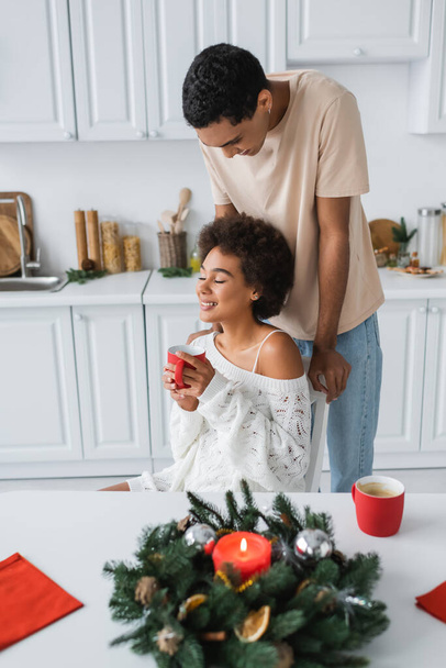 Африканская американка держит красную чашку и улыбается с закрытыми глазами рядом с молодым парнем и украшенный рождественский венок - Фото, изображение