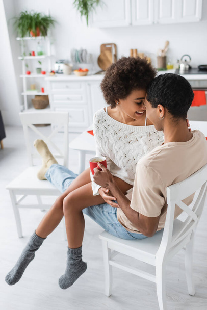 sexy african american woman in warm socks and white openwork sweater sitting on one chair with boyfriend in kitchen - Foto, afbeelding