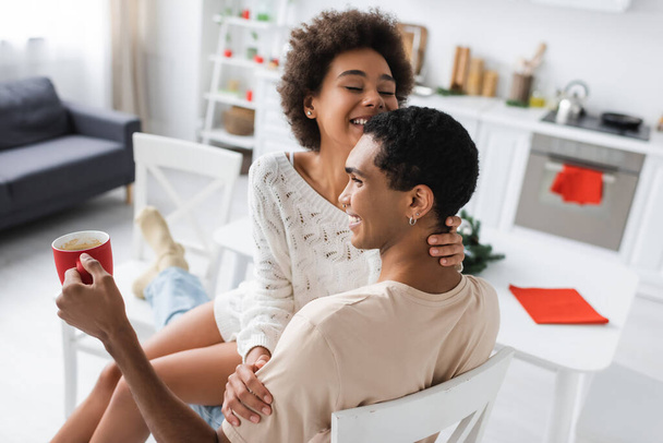 Mutfakta kucağında seksi kız arkadaşı otururken elinde kupa tutan mutlu Afro-Amerikan erkeği. - Fotoğraf, Görsel