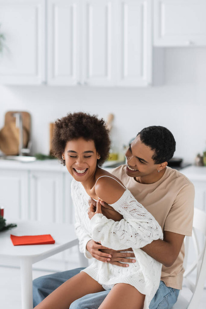 african american man sitting in kitchen and embracing laughing girlfriend in white openwork sweater - Φωτογραφία, εικόνα
