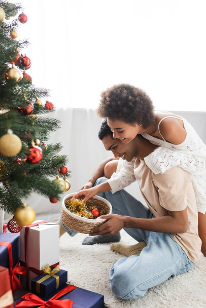 happy african american couple holding wicker basket with baubles near gift boxes and christmas tree - Foto, Imagem