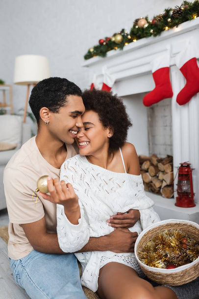 happy african american man embracing girlfriend holding christmas ball and tinsel in wicker basket - Foto, imagen