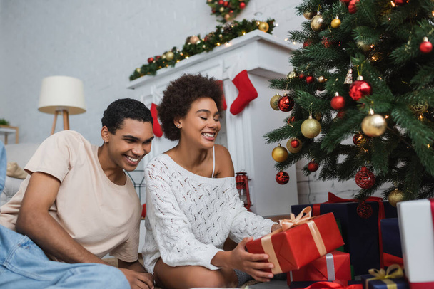 happy african american woman in white knitted sweater holding gift box near boyfriend  and christmas tree - Photo, Image