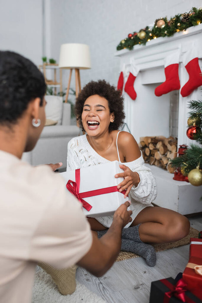 lachen Afrikaans amerikaanse vrouw houden geschenk doos terwijl zitten op de vloer in de buurt wazig vriendje - Foto, afbeelding