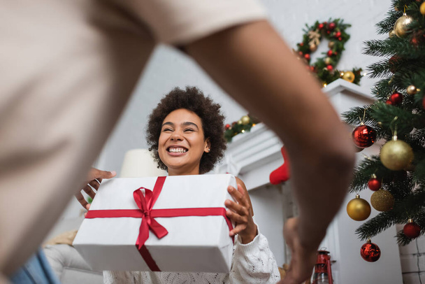 low angle view of overjoyed african american woman holding large gift box near christmas tree and blurred boyfriend - Zdjęcie, obraz