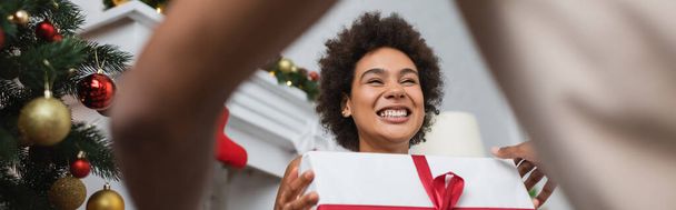 vista de ángulo bajo de la mujer afroamericana riendo con la Navidad presente cerca borrosa novio, bandera - Foto, imagen