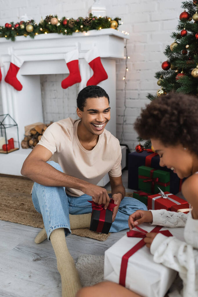 usmívající se africký Američan muž při pohledu na přítelkyně otevření dárkové krabice v blízkosti vánoční strom - Fotografie, Obrázek
