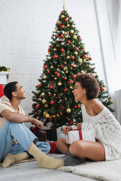 ευτυχισμένος αφρικανός Αμερικανός άνδρας κοιτάζοντας το χριστουγεννιάτικο δέντρο κοντά σέξι φίλη με κουτί δώρου - Φωτογραφία, εικόνα